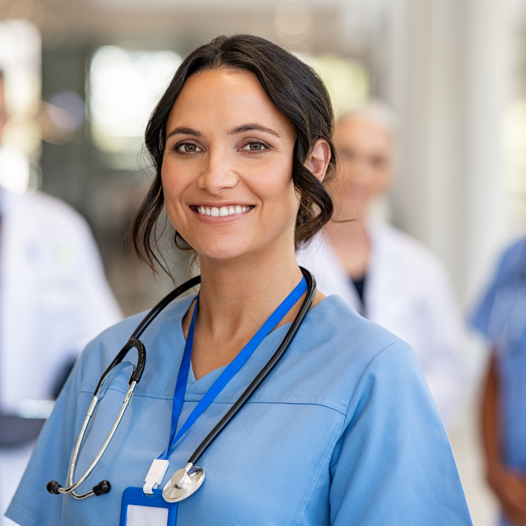 Woman in blue scrubs with a stethoscope and name badge, smiling in a hospital corridor with colleagues in the background.
