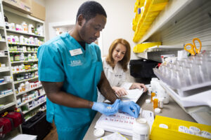 A pharmacy student prepares medication as a pharmacist oversees and reviews paperwork in a pharmacy workspace.
