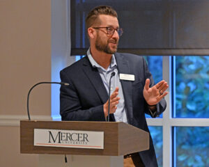 A man in a suit speaks at a podium labeled Mercer University in a well-lit room with large windows.