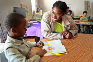 An adult and a child sit at a table in a classroom, reading a picture book together.