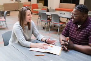 Two people sit at a table talking, with an open book and pen between them in a modern, casual setting.