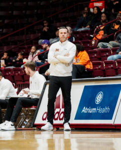 Mercer men's basketball coach Ryan Ridder stands with his arms crossed on the sidelines during a game.
