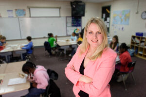 A woman in a pink blazer stands smiling in a classroom with children working at desks in the background.