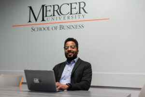 A man in a collared shirt and blazer sits at a laptop in front of a wall, which reads, Mercer University School of Business.