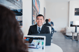 Two people wearing business attire sit at a table with laptops and talk with each other.