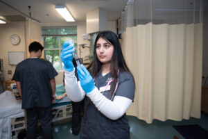 A Mercer nursing student in scrubs and gloves prepares a syringe in a hospital room, with another person in the background.