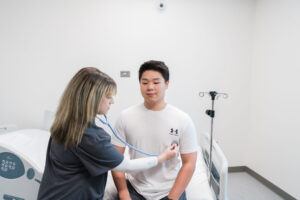 A nursing student uses a stethoscope to examine a seated patient in a medical exam room.