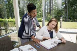 A woman stands beside a seated woman who is reading a book at a table near large windows overlooking greenery.