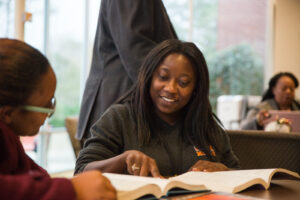 Two women sit at a table, looking at an open book together in a study area with people in the background.