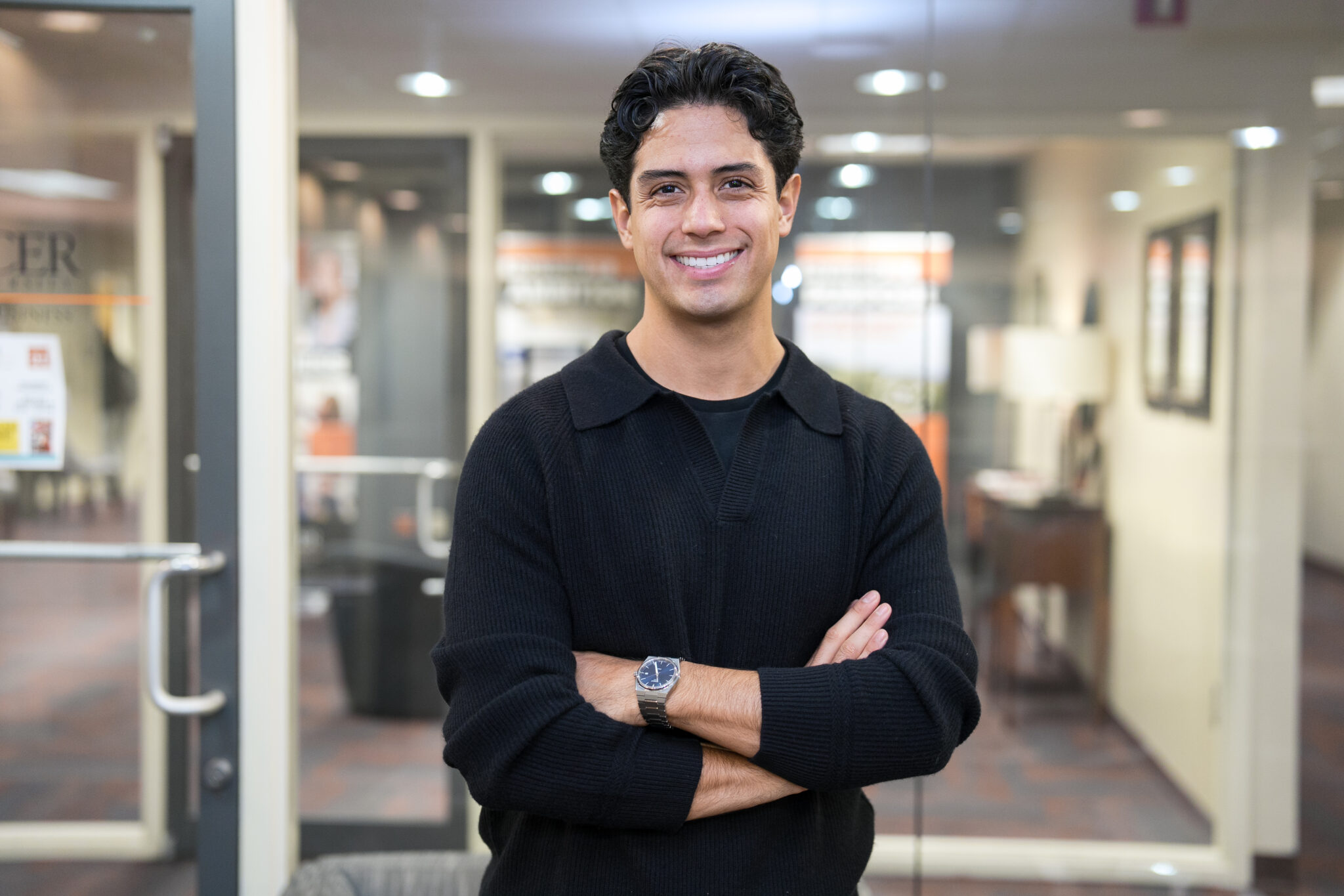 A person with dark hair in a black sweater stands indoors with arms crossed, smiling at the camera.