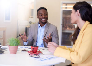 Two professionals engaged in a discussion at a meeting table. One person, in a tan blazer, is smiling and gesturing with hands open, while the other, partially visible, in a yellow blazer, appears to be listening attentively. The table is scattered with documents, digital devices, and cups, suggesting a business meeting environment.