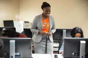 A person wearing an orange Mercer University T-shirt under a gray suit jacket smiles while standing in an office-like classroom setting with multiple students working on desktop computers.