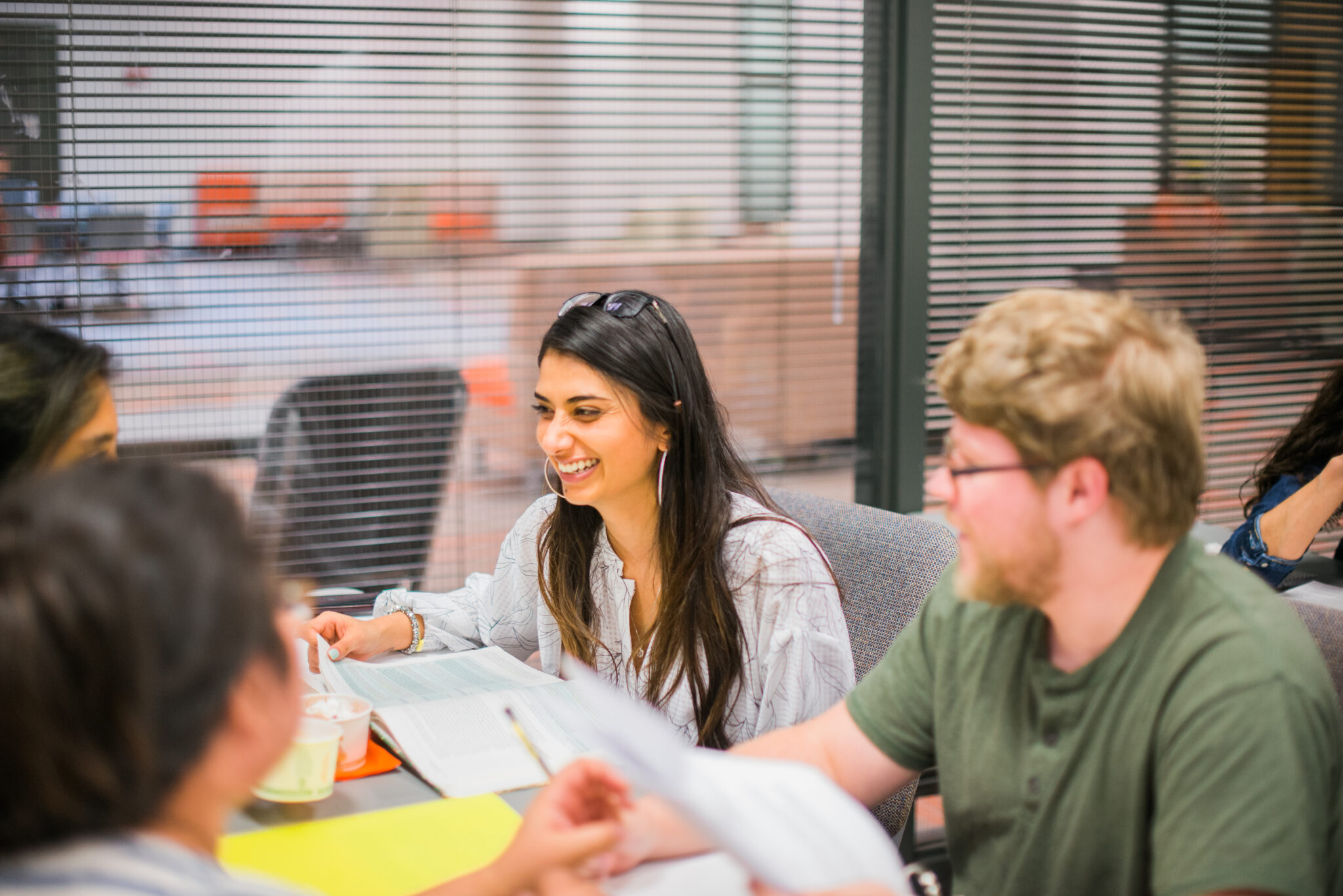 Four people sit at a table with books and papers, having a discussion in a study area with glass walls.
