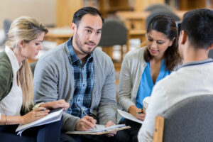 Four adults sit in a circle having a discussion, with notebooks, in a library or meeting room.