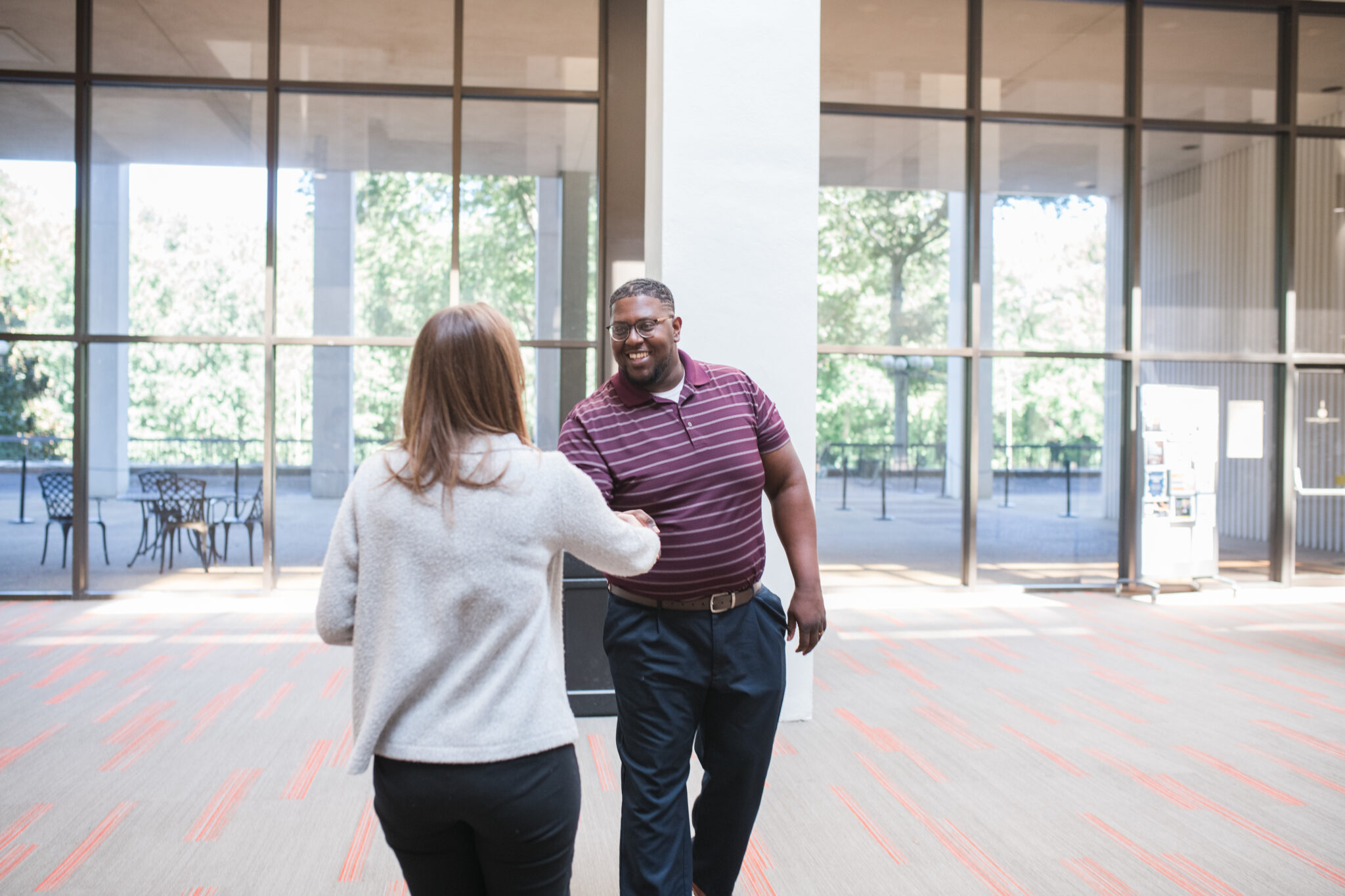 Two people shake hands in a spacious, sunlit lobby with large windows and outdoor seating visible in the background.