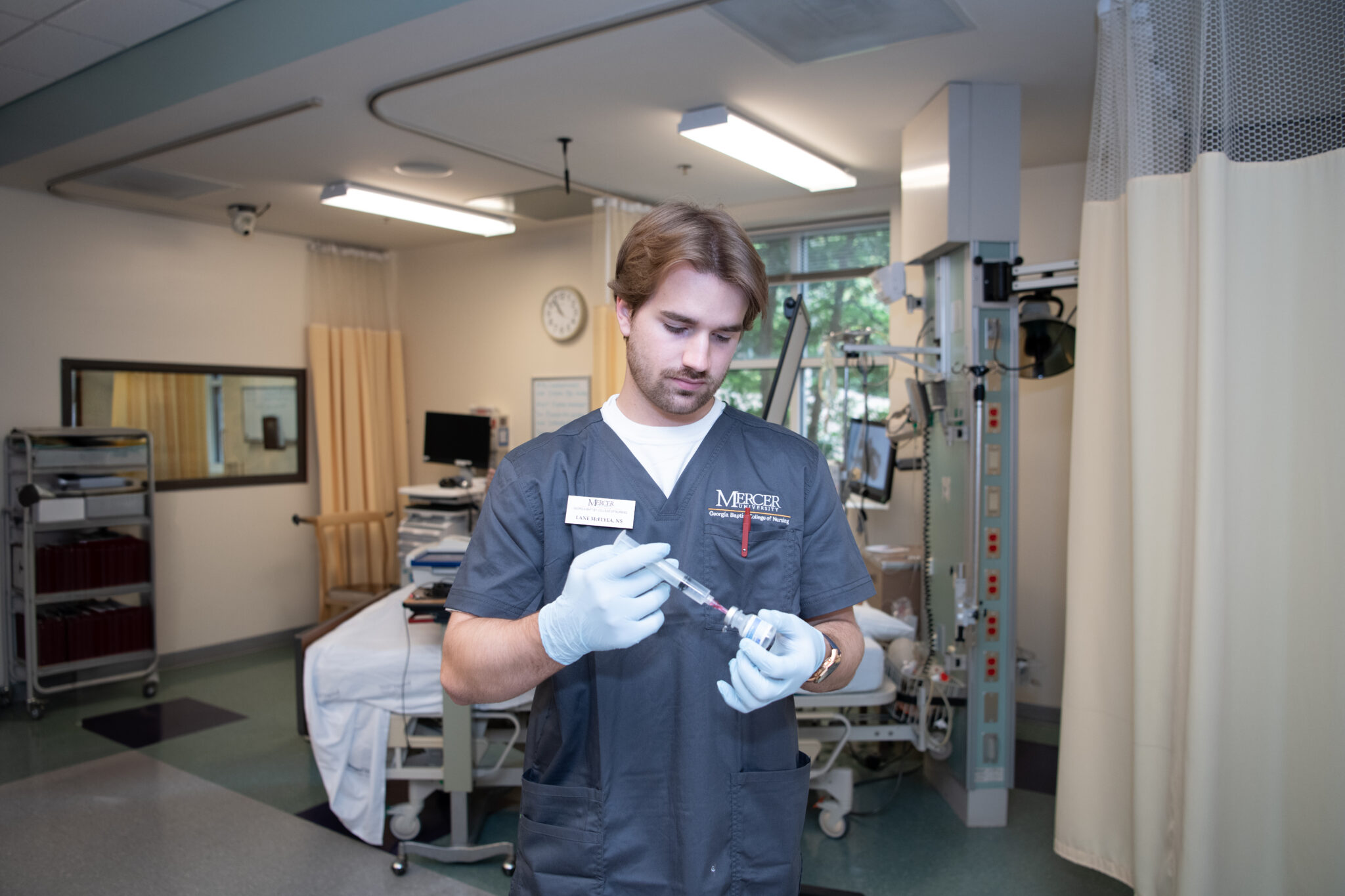 A Mercer nursing student in scrubs prepares a syringe in a hospital room with medical equipment in the background.