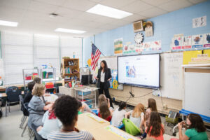 A teacher stands at the front of a classroom as students sit at tables, facing her and a screen displaying a lesson.