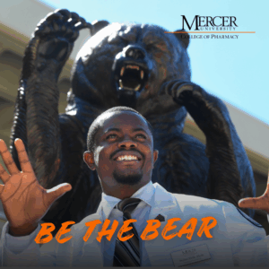 A man in a white coat poses with raised hands in front of a large bear statue at Mercer University College of Pharmacy.