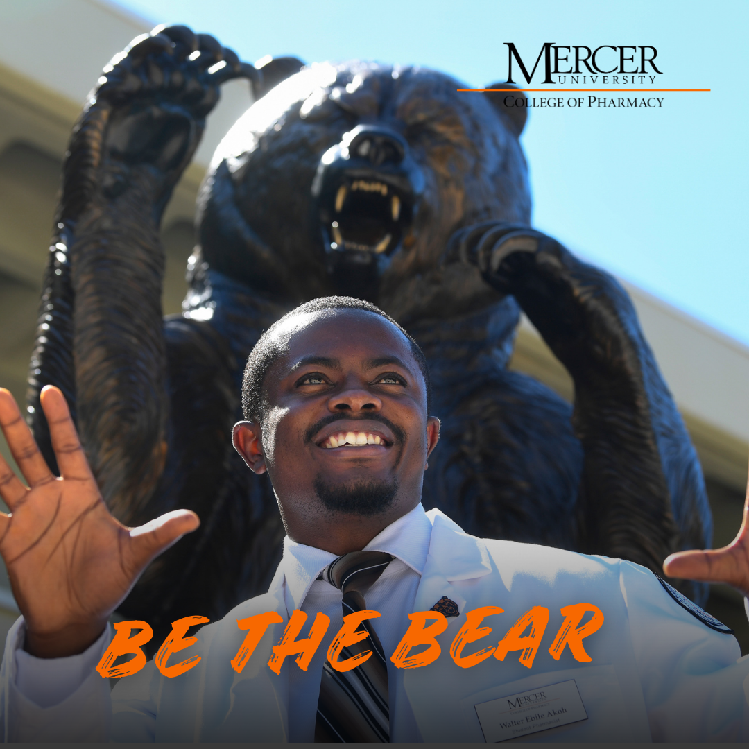 A man in a white coat poses with raised hands in front of a large bear statue at Mercer University College of Pharmacy.