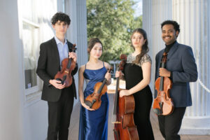 Four formally dressed young adults stand outside holding string instruments including violins, viola, and cello.