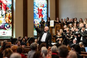 A choral director leads an audience in song while a choir sings behind him.