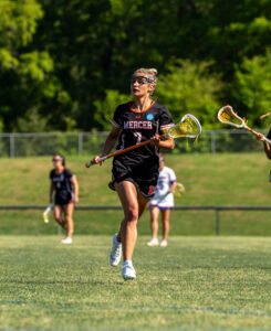 A Mercer lacrosse player runs on a grassy field holding a lacrosse stick during a game.
