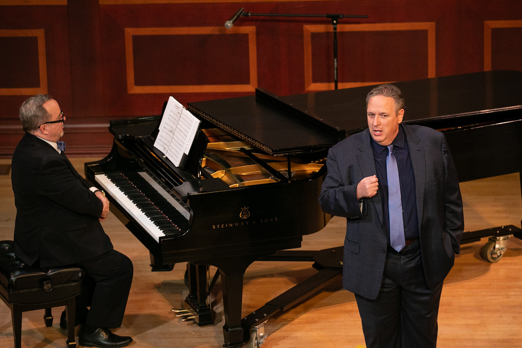 A man in a suit sings while another man sits at a grand piano with sheet music on stage.