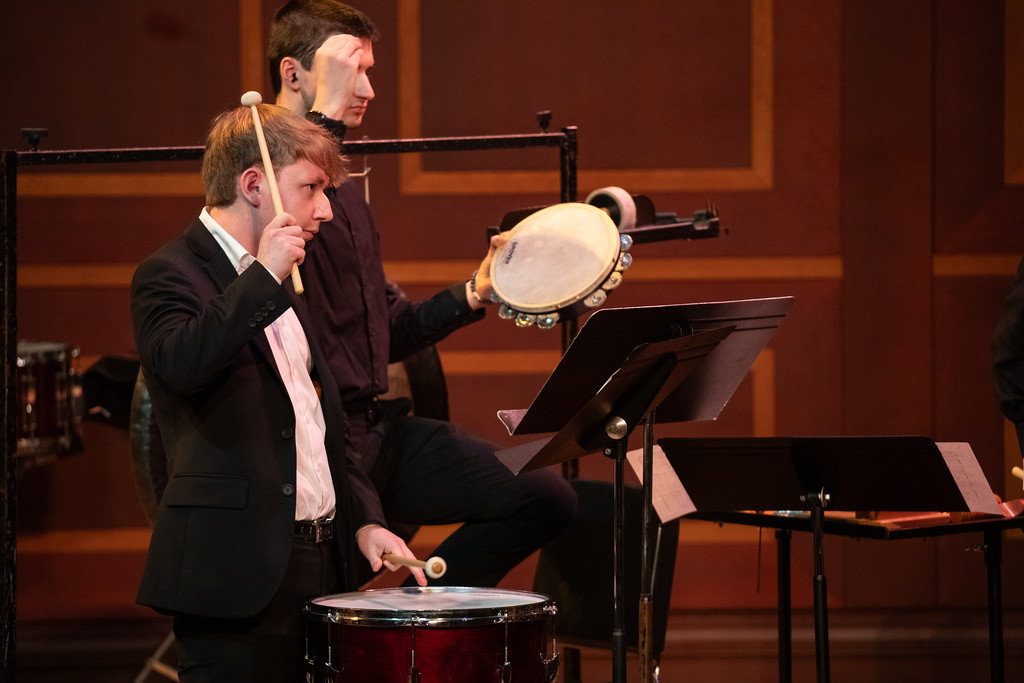 Two musicians perform on stage, one playing a drum and the other holding a tambourine.