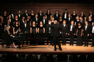 A choir performing on stage with a conductor in the foreground and a pianist seated at a piano to the left.