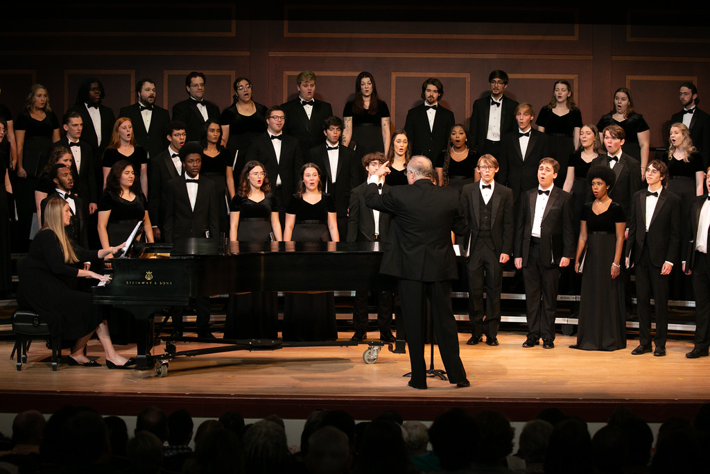 A choir performing on stage with a conductor in the foreground and a pianist seated at a piano to the left.