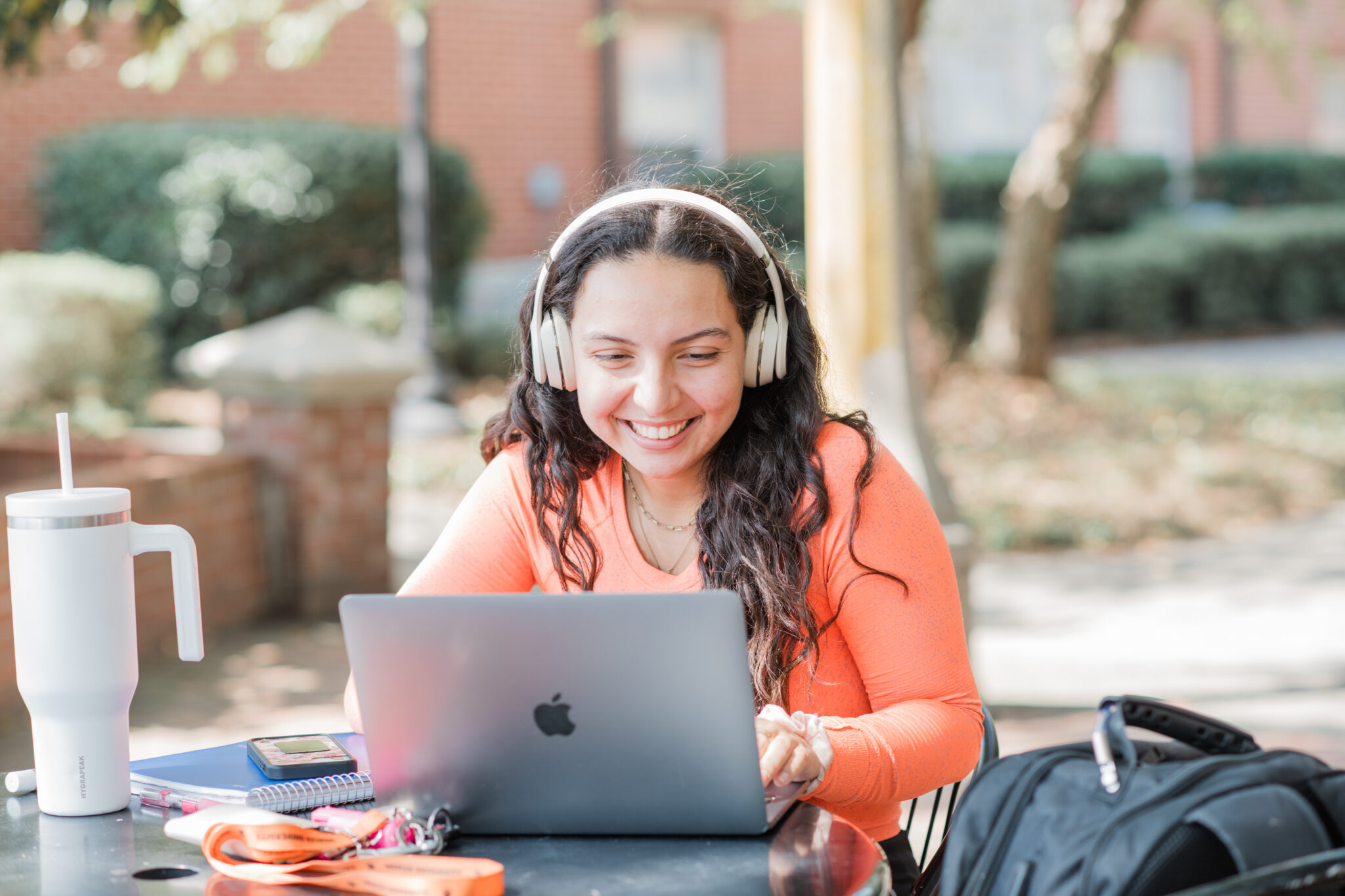 Woman wearing headphones smiles while using a laptop at an outdoor table with notebooks and a water bottle.
