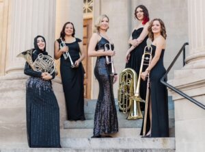 Five women in black formal dresses hold brass instruments, including the french horn, trumpet, tuba and trombone.