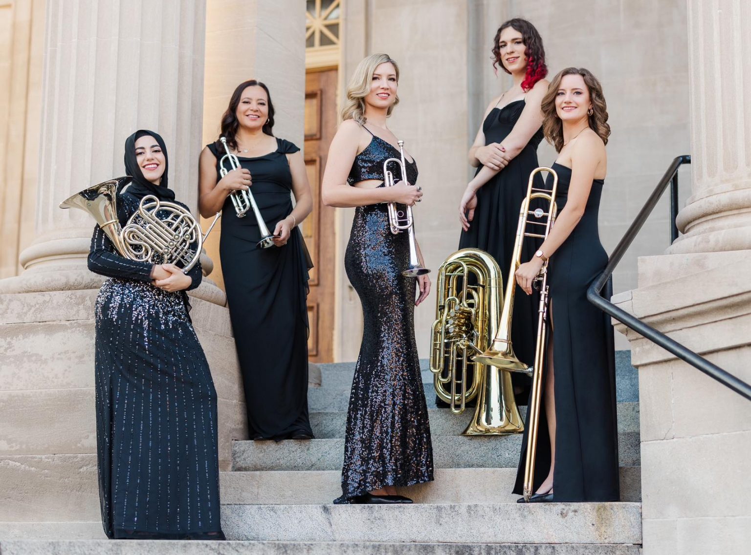 Five women in black formal dresses hold brass instruments, including the french horn, trumpet, tuba and trombone.