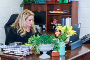 A woman sits behind a desk topped with a phone, computer and plants.
