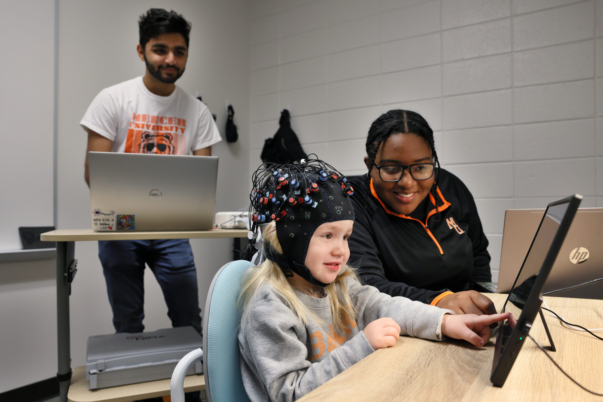 A Mercer student stands looking at a computer, while another student plays with a child on the floor. The child is wearing a cap fitted with technology.