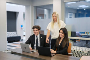 Three people in business attire work on laptops at a table in a modern office space, with one person standing and pointing.