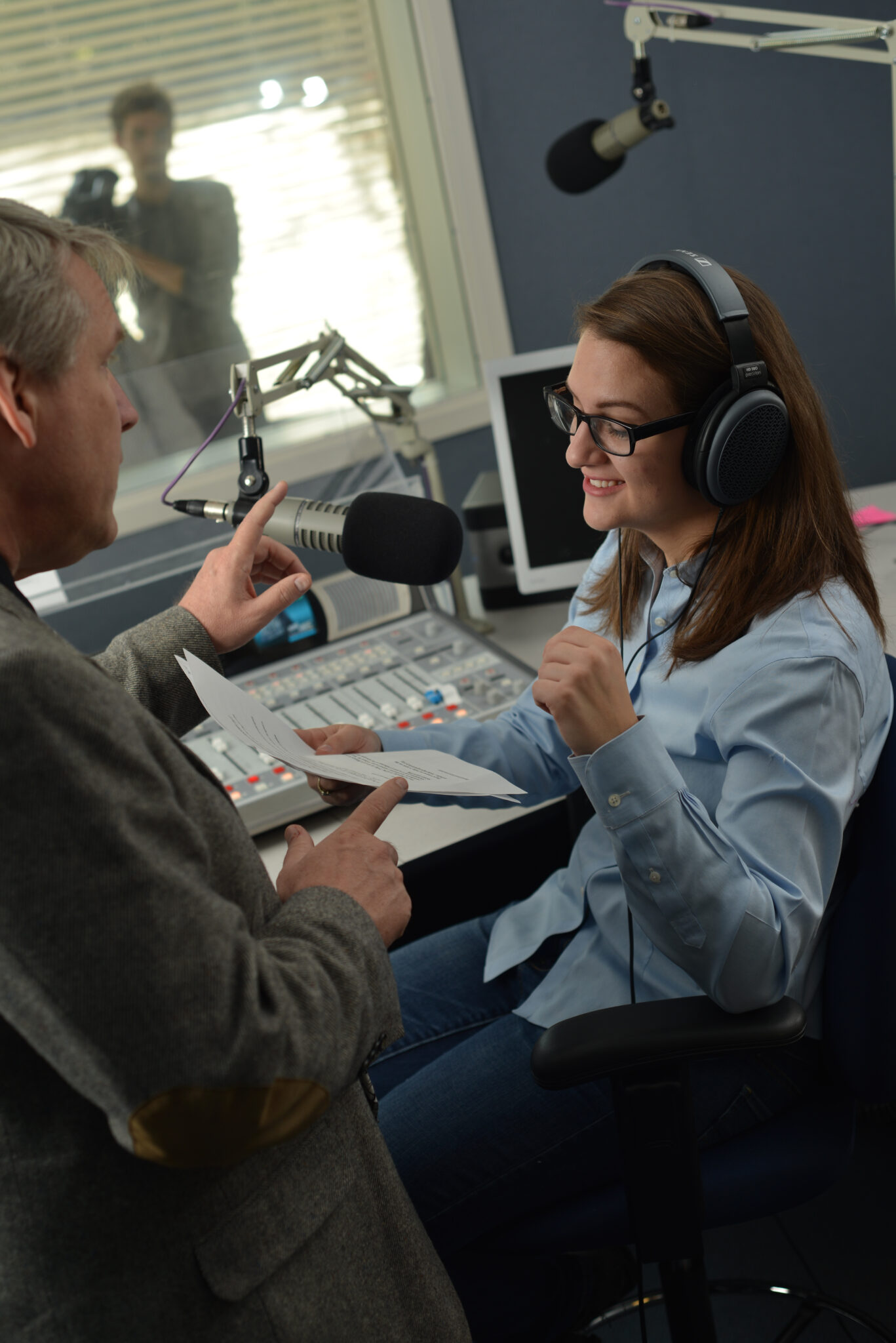 A Mercer student wearing headphones sits at a radio studio desk, talking with a man while recording.