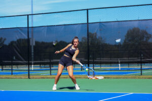 Mercer women's tennis player prepares to hit a forehand shot on an outdoor hard court, with a chain-link fence in the background.