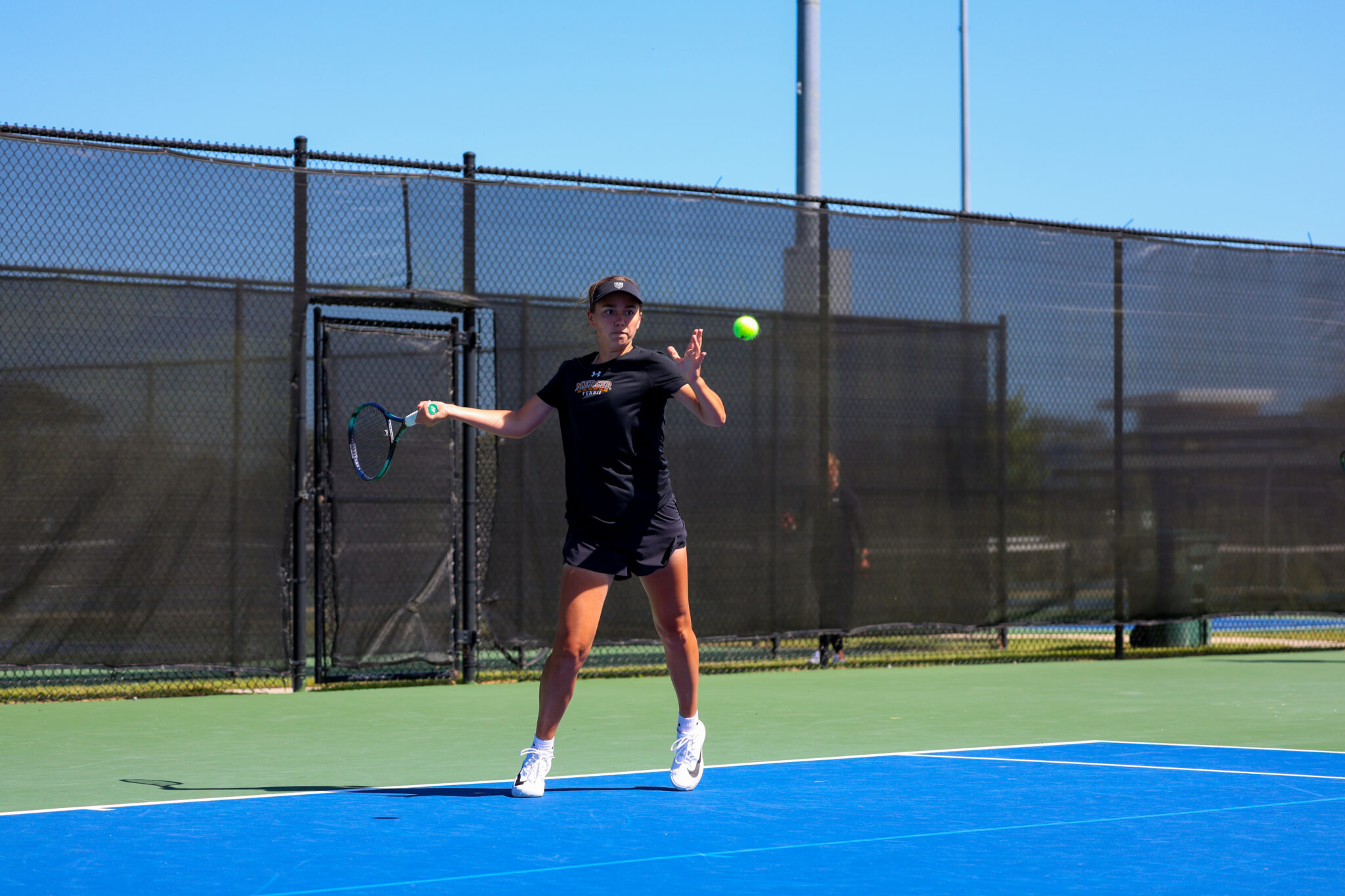 Mercer women's tennis player prepares to hit a tennis ball with a racket on an outdoor court under clear skies.