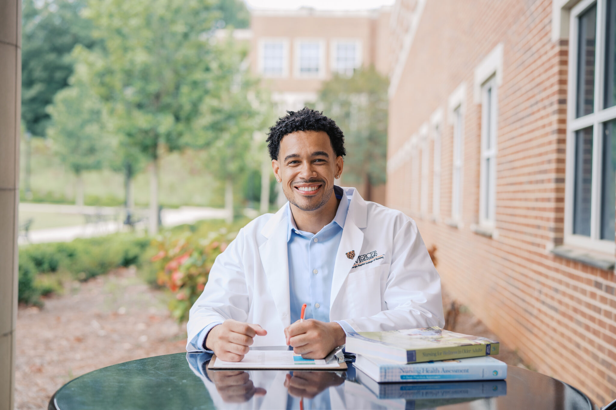 A nursing student in a white coat sits at an outdoor table with books and a clipboard, smiling at the camera.
