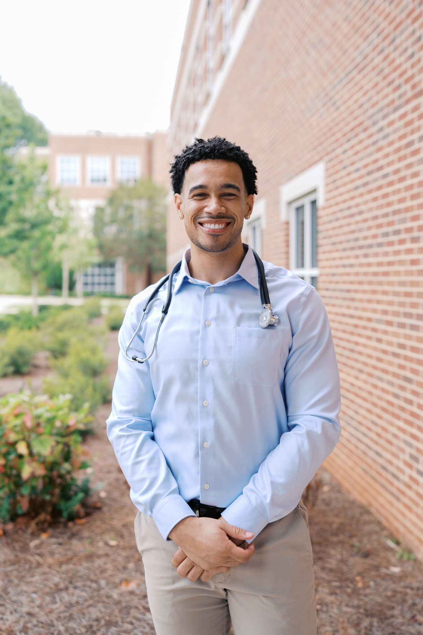 A man wearing a light blue shirt and stethoscope stands outside a brick building, smiling at the camera.