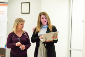 Two women stand indoors, one holding a laptop and speaking, the other listening.