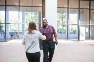 Two people shake hands in a spacious, sunlit lobby with large windows and outdoor seating visible in the background.