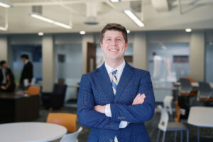 A Mercer student wearing a business suit stands smiling with his arms crossed in a classroom.