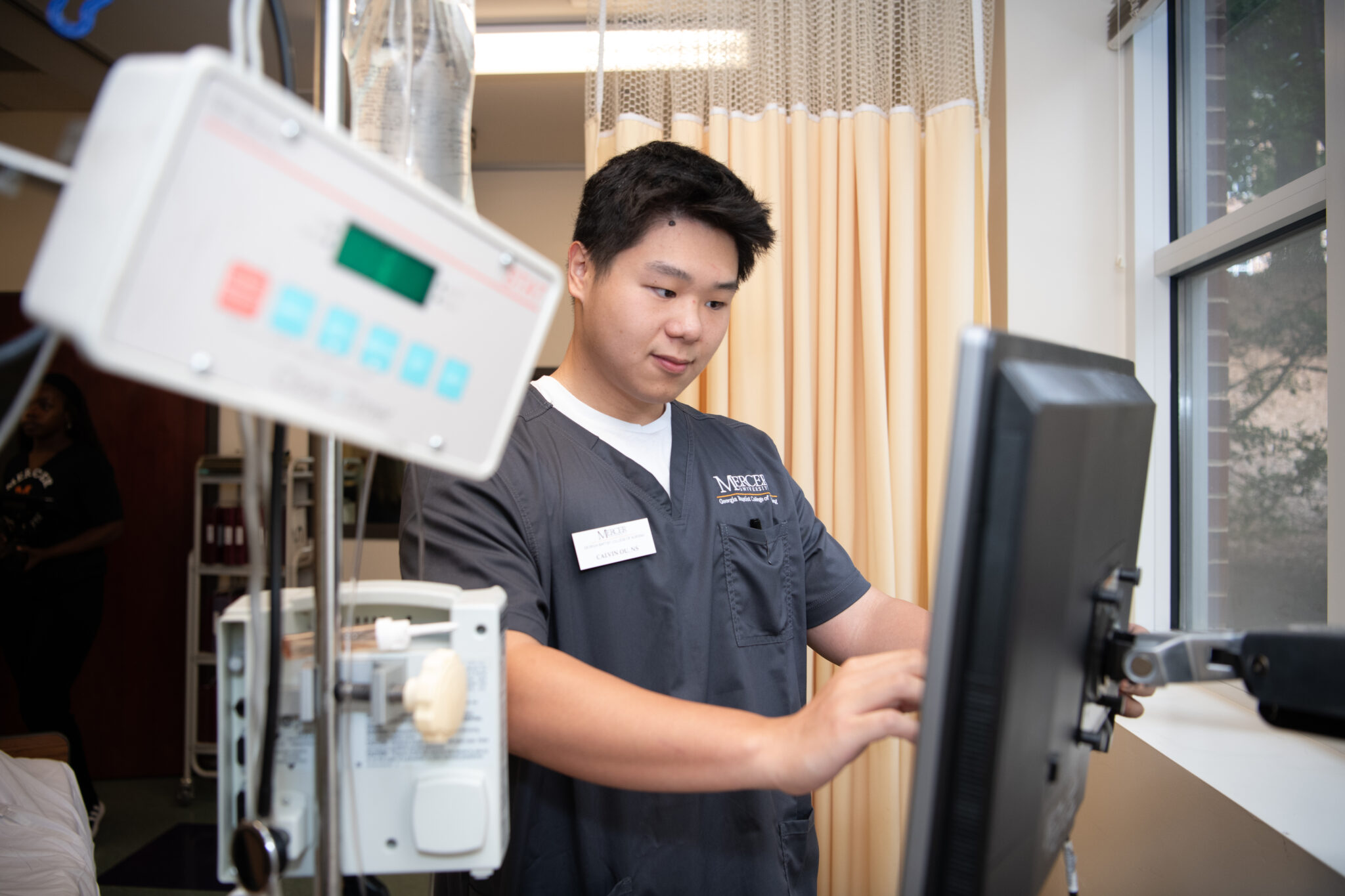 A Mercer nursing student uses a computer in a hospital room with medical equipment nearby.