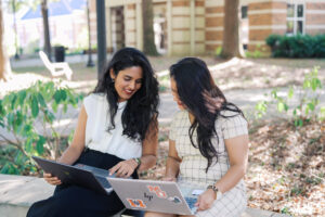 Two Mercer students in business attire hold their laptops while sitting on a ledge outside.