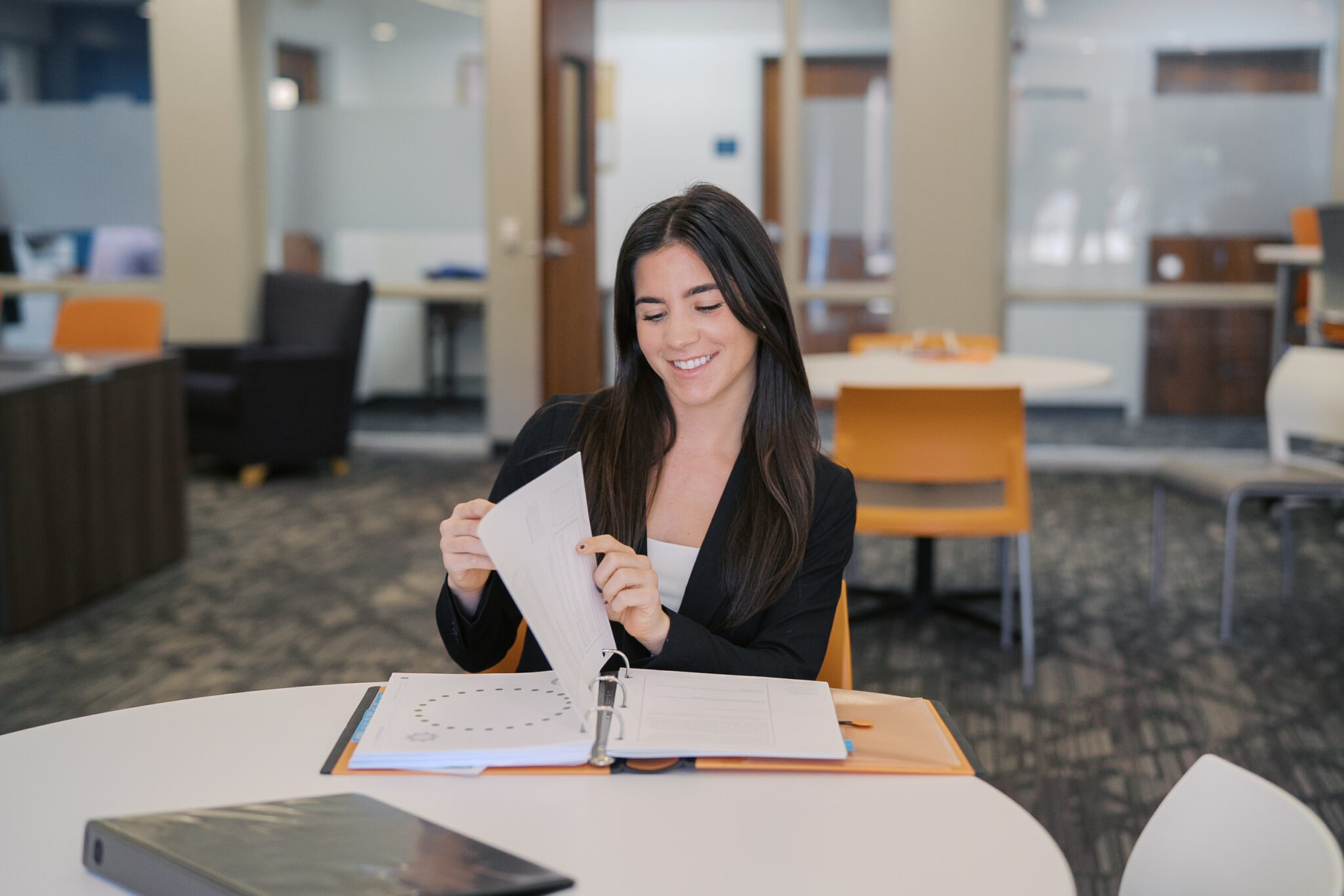 A woman sitting at a round table in an office looks through papers in a binder and smiles.