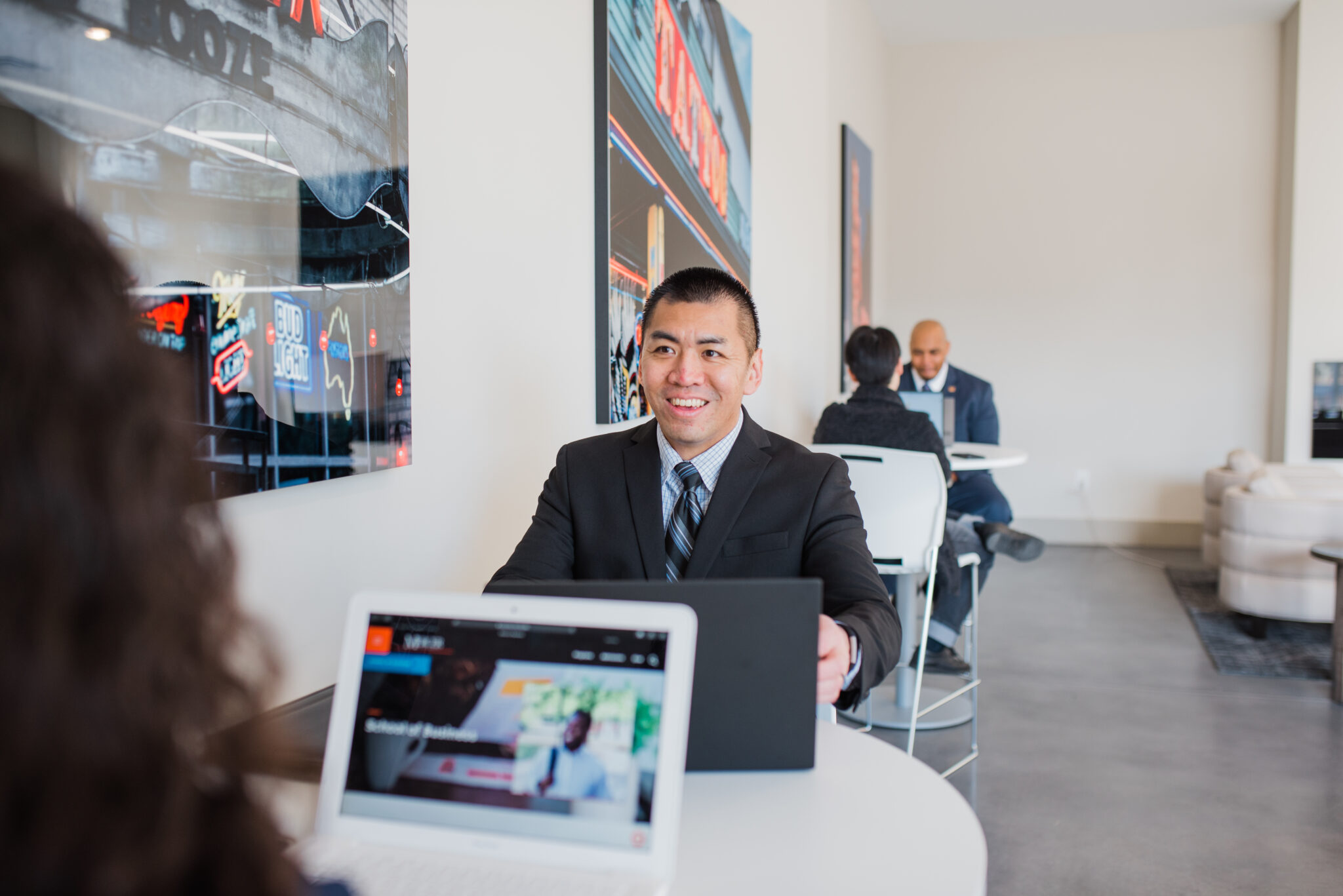 Man in a suit smiling while working on a laptop at a table in a modern, bright area with other people in the background.