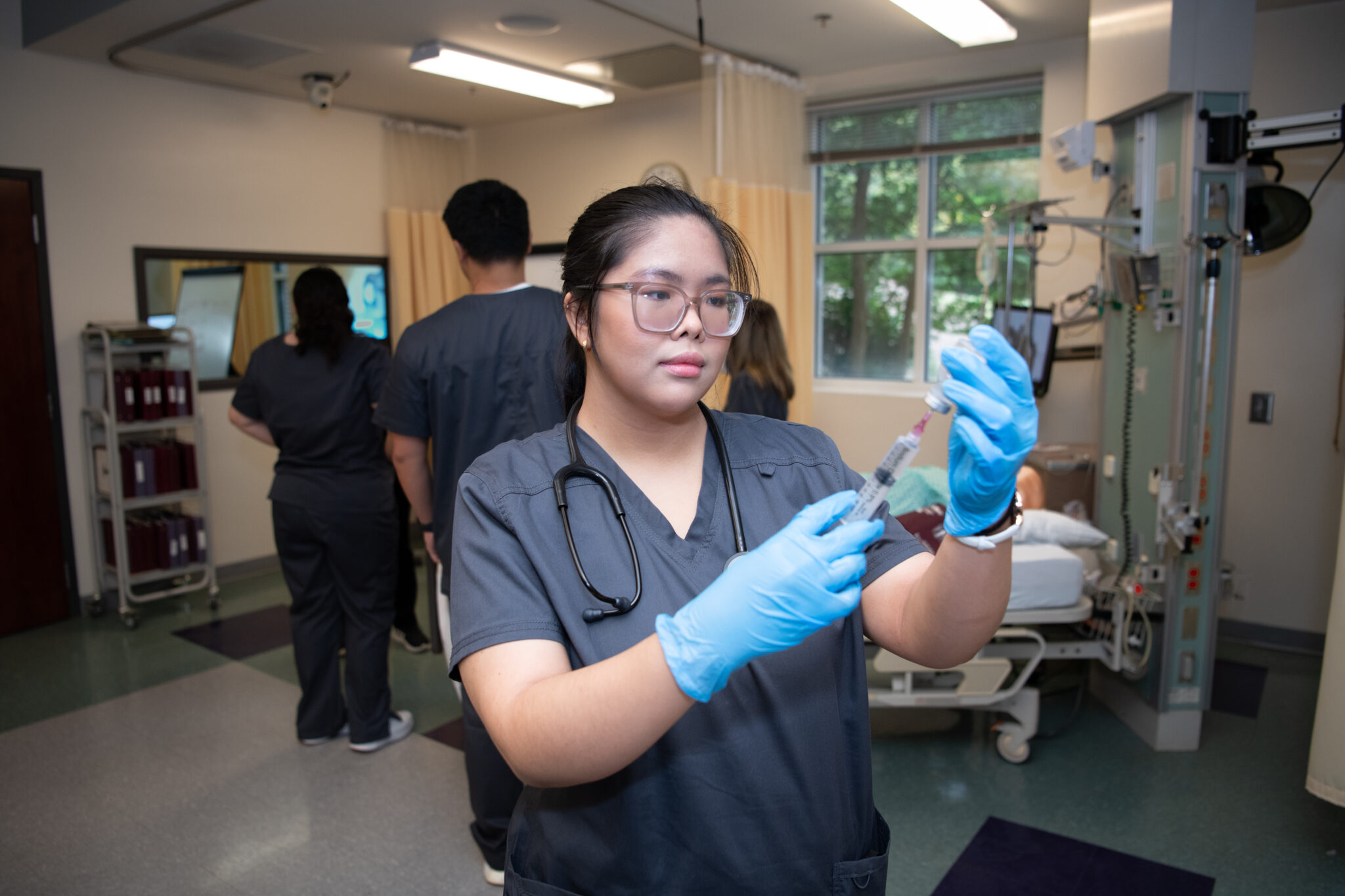 A Mercer nursing student in scrubs and gloves prepares a syringe in a simulated hospital room.