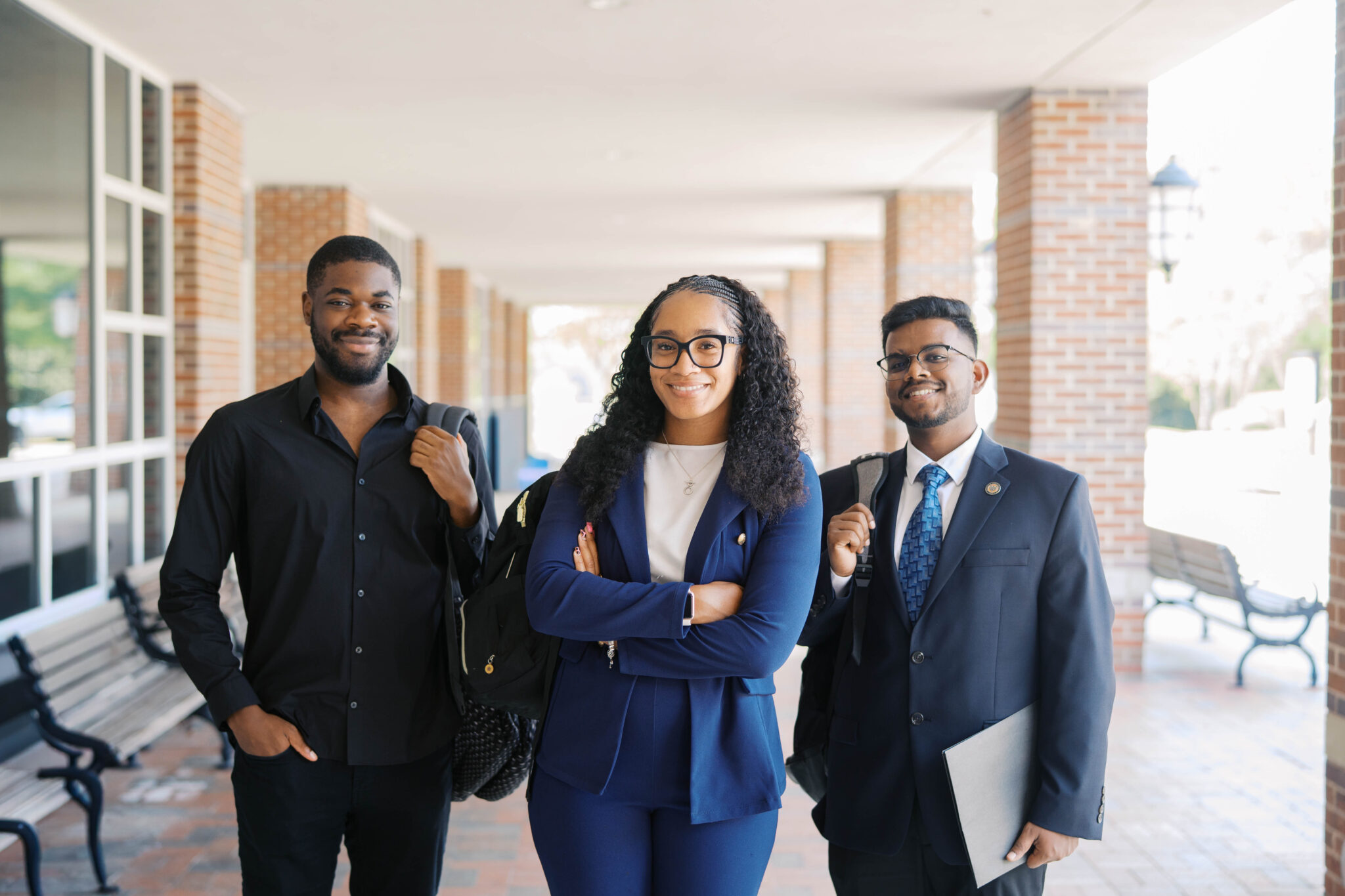Three professionally dressed Mercer students stand together in a covered outdoor walkway, smiling at the camera.